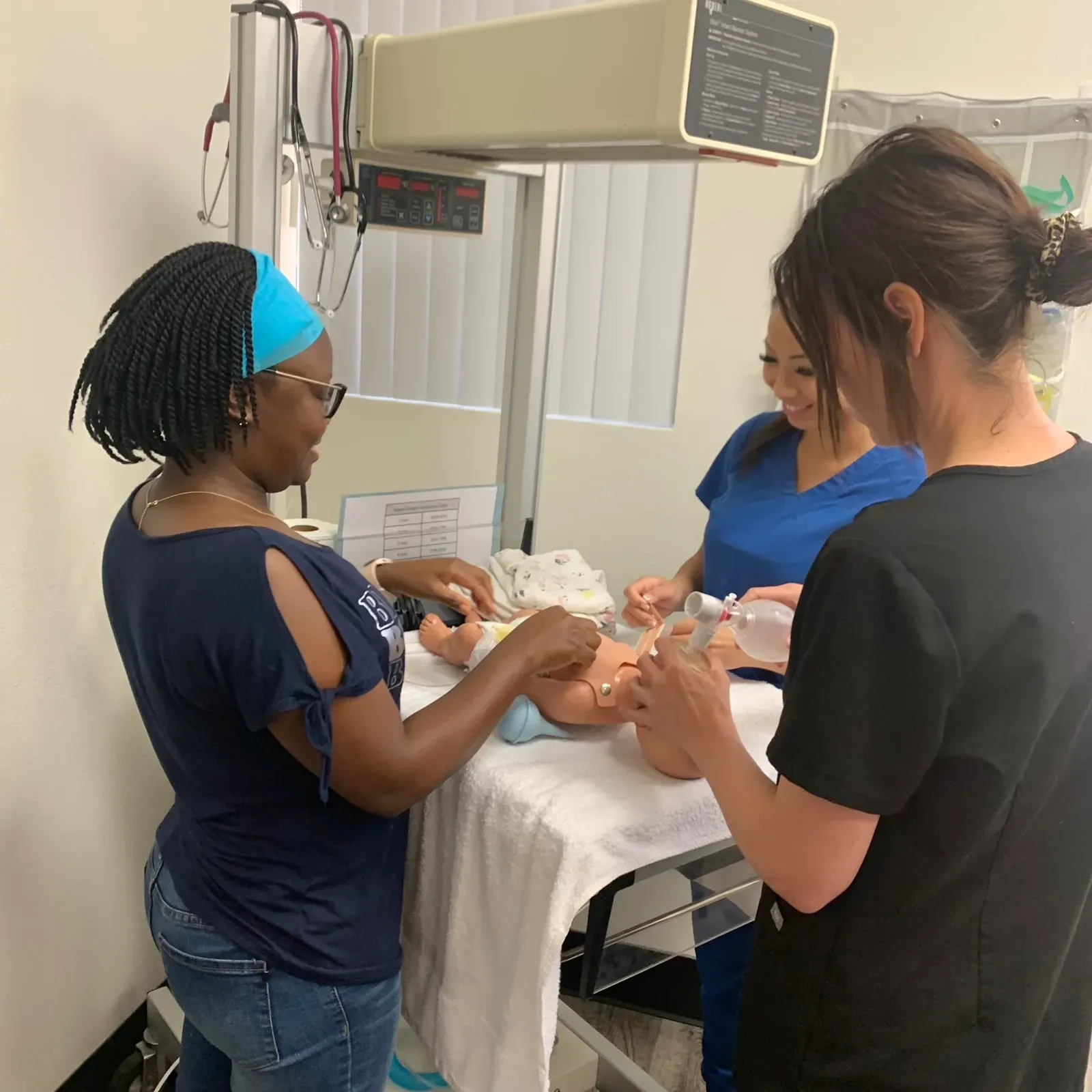 Three students working a newborn manikin with a bag-valve-mask under a radiant warmer during NRP Megacode skills training at Heartbeat Haven