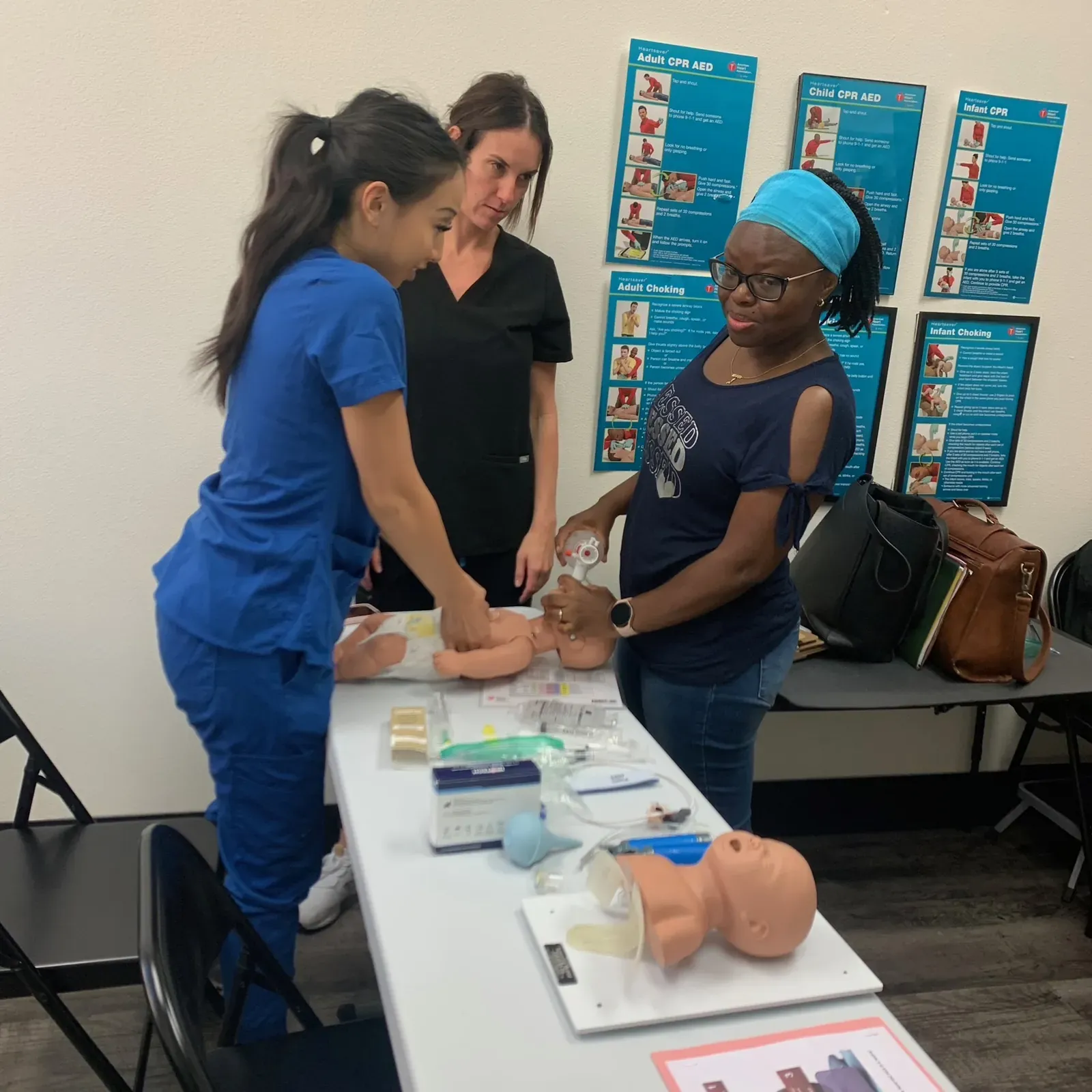 Adriana Park and two students working an infant CPR manikin with a bag-valve-mask during a pediatric resuscitation skills station at Heartbeat Haven, AHA CPR posters on the wall behind them